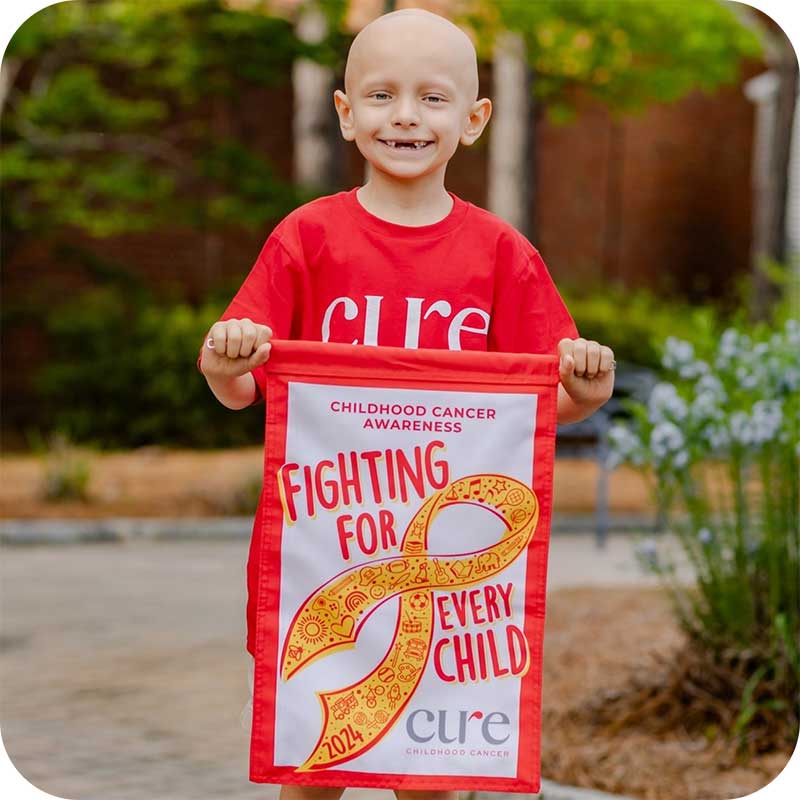 Smiling child in red CURE shirt holding orange and gold ribbon sign