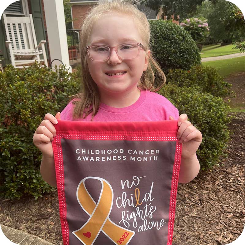 Smiling child in pink CURE shirt with awareness flag featuring gold ribbon